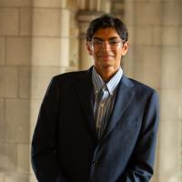 a person wearing a suit and glasses standing in a stone hallway and smiling